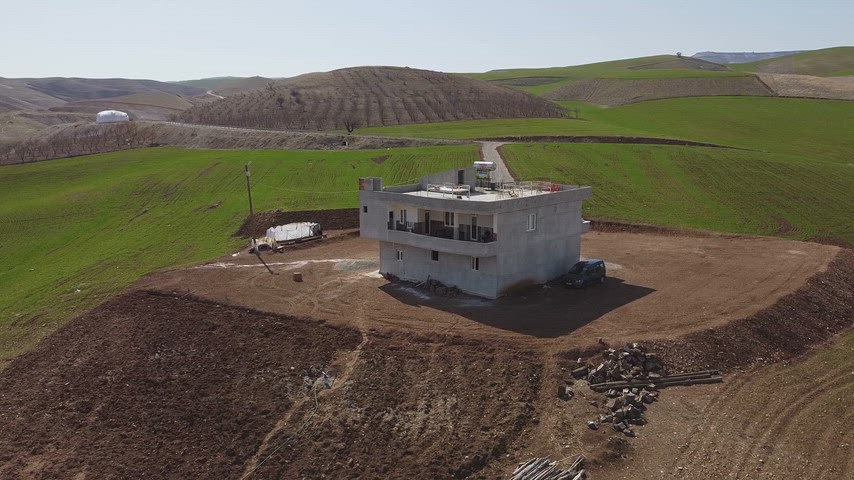 Drone ile havadan çekilmiş, yeşil ekili tarlalar, toprak yol ve dalgalı tepeler arasında yer alan ev manzarası.