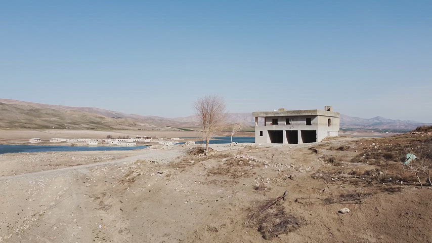 Kurak arazi üzerinde bulunan beton evin, sakin su kıyısı ve uzak dağlarla çevrili geniş doğal manzarası.