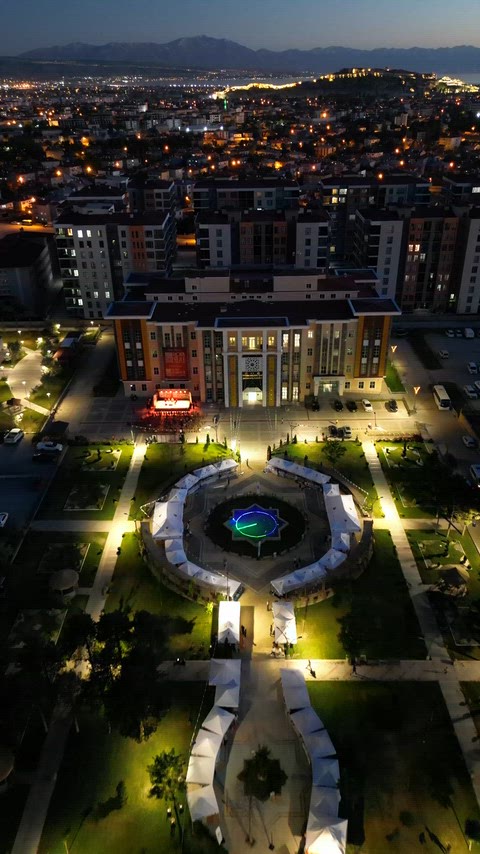 Van'ın Tuşba ilçesinde bulunan belediye hizmet binasının akşam saatlerinde havadan görünümü. Aydınlatılmış bina, çevredeki yerleşim alanları ve uzakta görülen dağ silsilesi etkileyici bir şehir manzarası oluşturuyor.