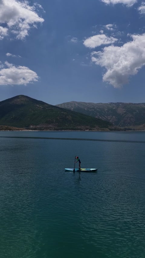 SUP yapan sporcunun drone görüntüsü, berrak göl suları, dağ manzarası ve doğal kıyı peyzajını bir araya getiriyor.